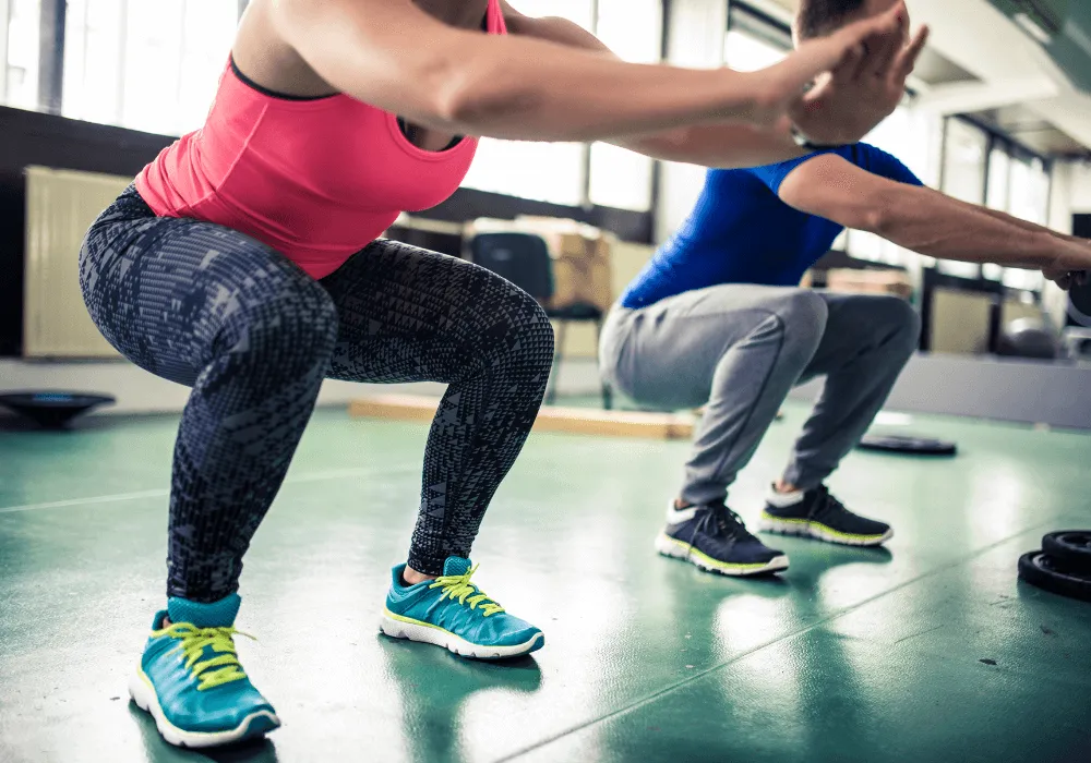 Two individuals performing squats in a gym, dressed in athletic wear. The woman wears a pink tank top and patterned leggings, the man a blue shirt.