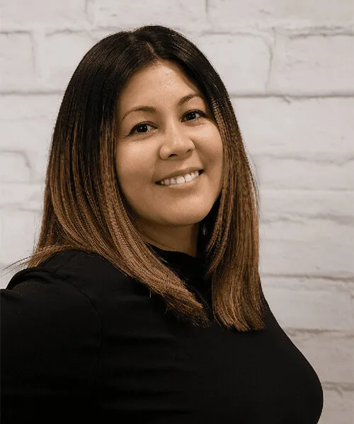 A woman with straight, shoulder-length hair stands against a textured white brick wall, wearing a black top.